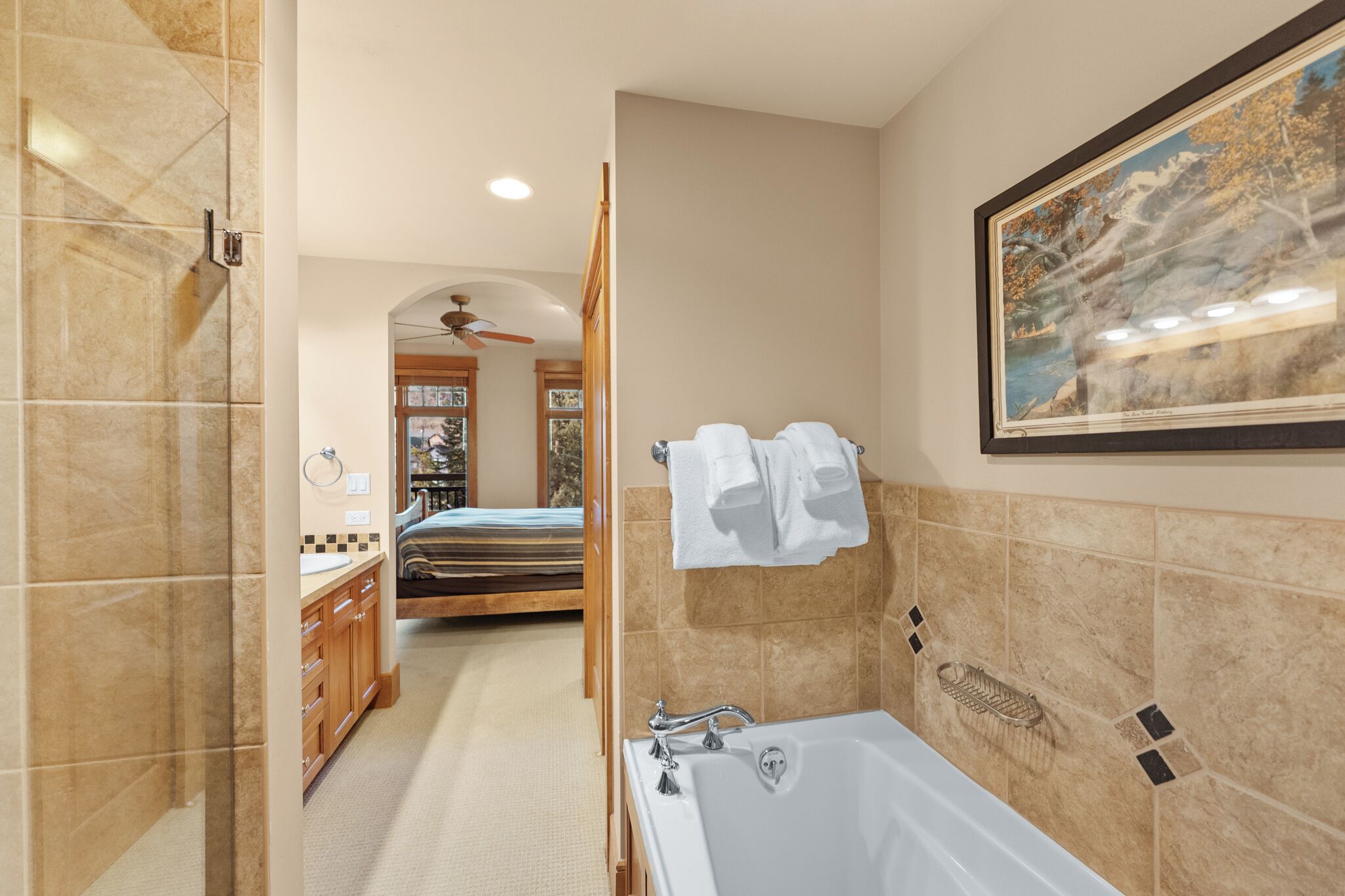 A modern bathroom with a bathtub, shower, and vanity. There is a framed picture on the wall and a door leading to another room.