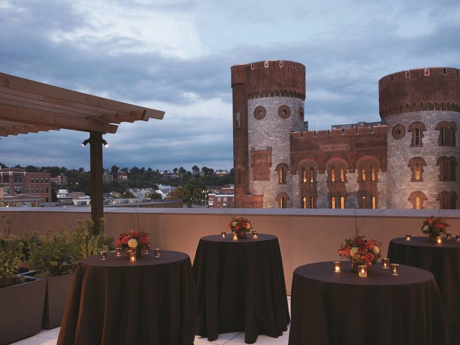 Eine Dachterrasse mit runden Tischen, gedeckt für ein Essen, mit Blick auf ein historisches Gebäude mit zwei markanten Türmen.