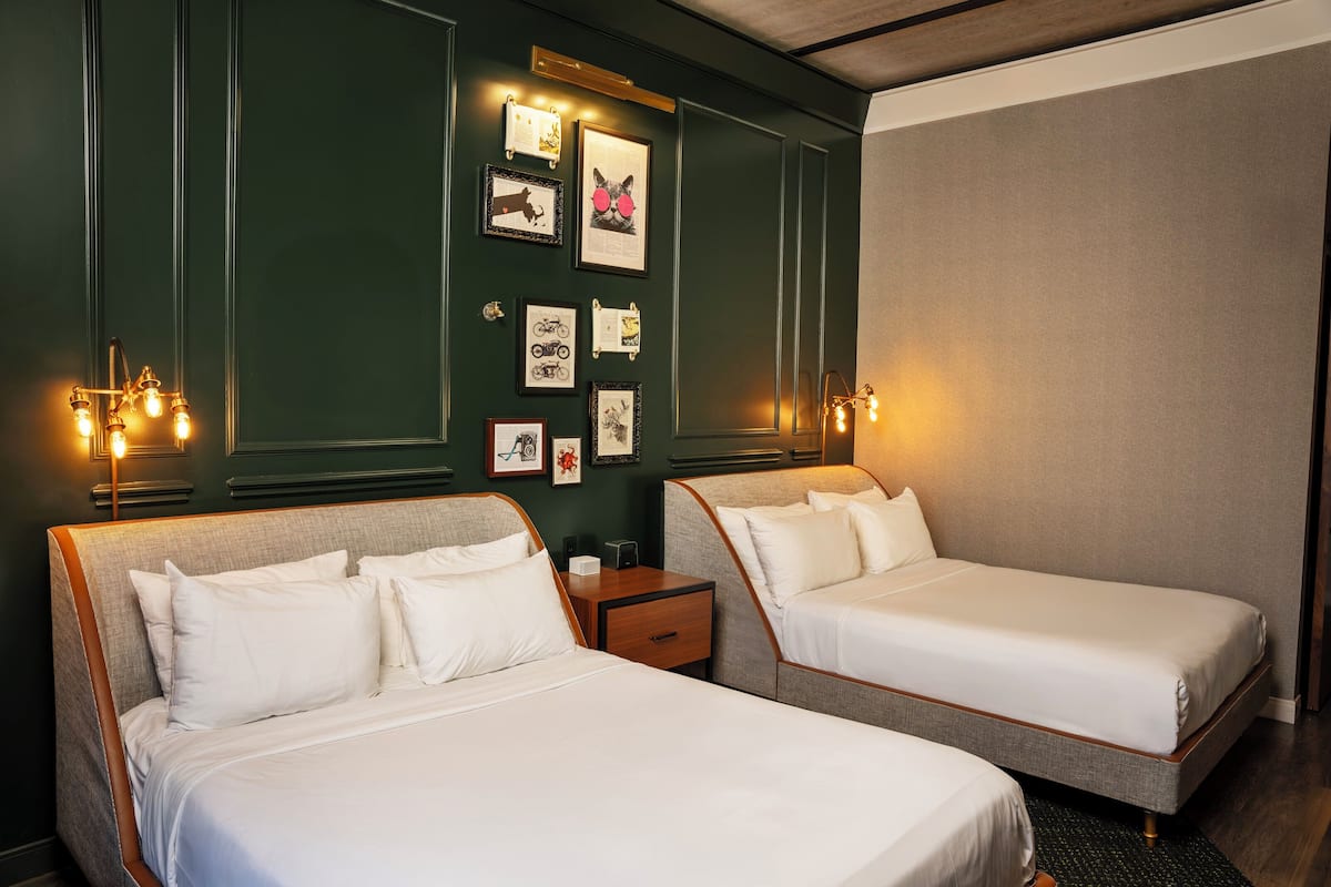 A hotel room with two beds, a nightstand, and wall-mounted lighting.