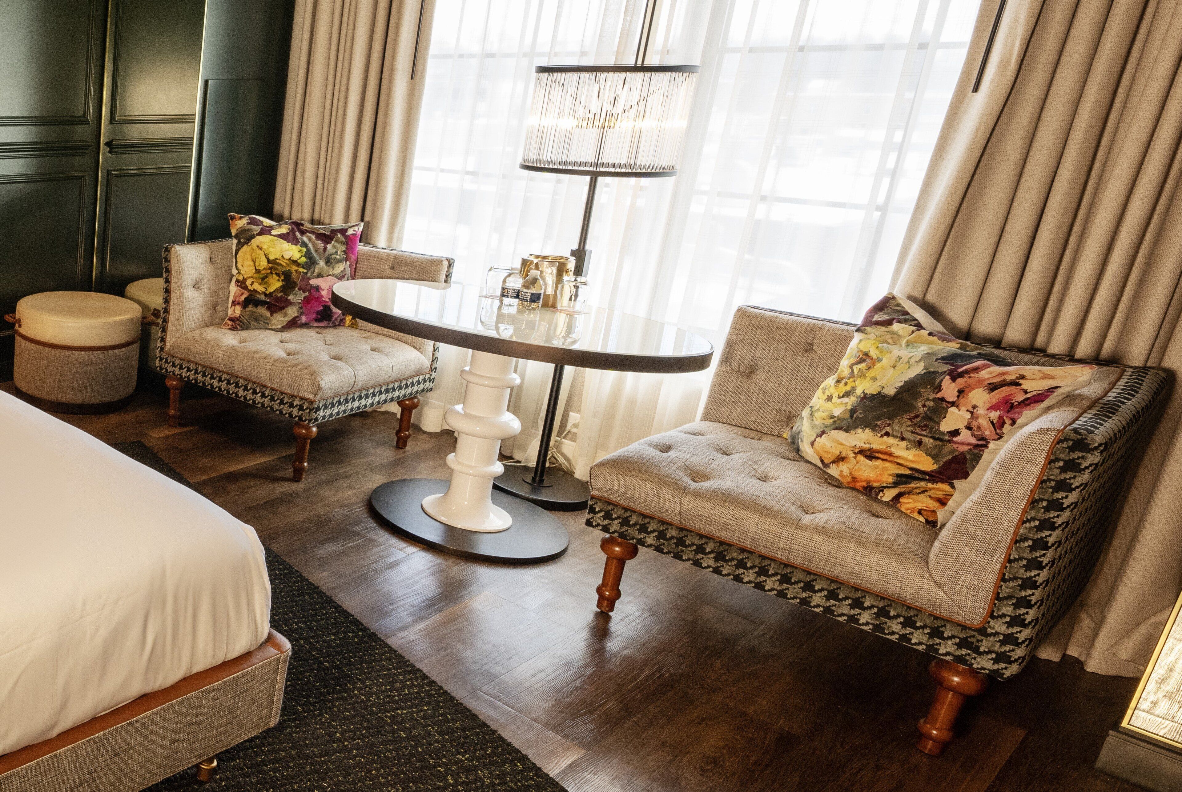Une chambre d’hôtel comprenant un lit, deux fauteuils, une table ronde, une lampe sur pied et une fenêtre avec des rideaux.