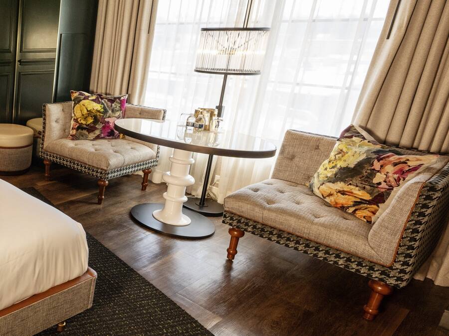 Une chambre d’hôtel comprenant un lit, deux fauteuils, une table ronde, une lampe sur pied et une fenêtre avec des rideaux.
