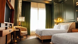 A hotel room with two beds, a desk, a chair, a lamp, and a large window with curtains.