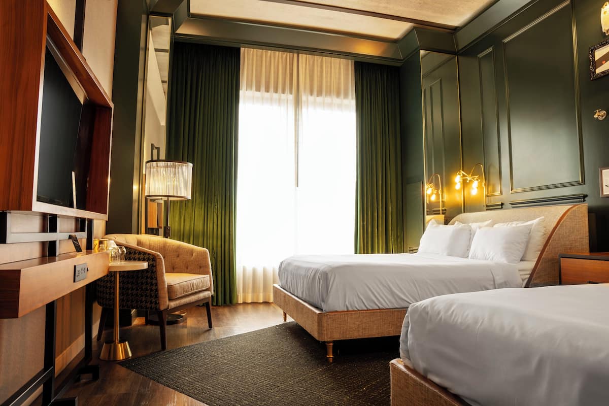 A hotel room with two beds, a desk, a chair, a lamp, and a large window with curtains.