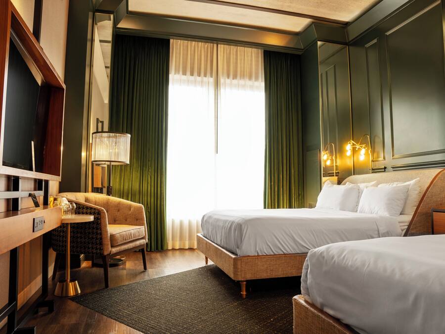 A hotel room with two beds, a desk, a chair, a lamp, and a large window with curtains.