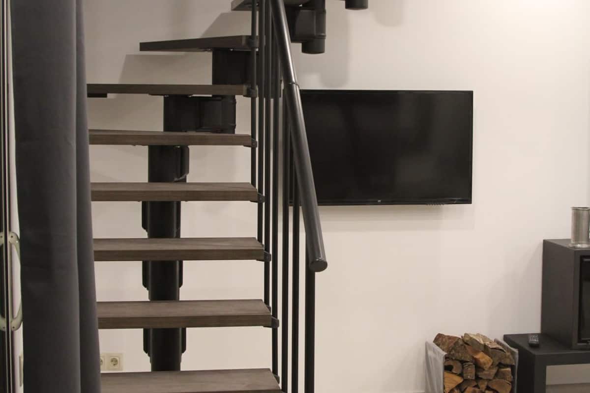 Una escalera moderna con barandilla negra, un televisor de pantalla plana montado en la pared y una pila de leña en el suelo.