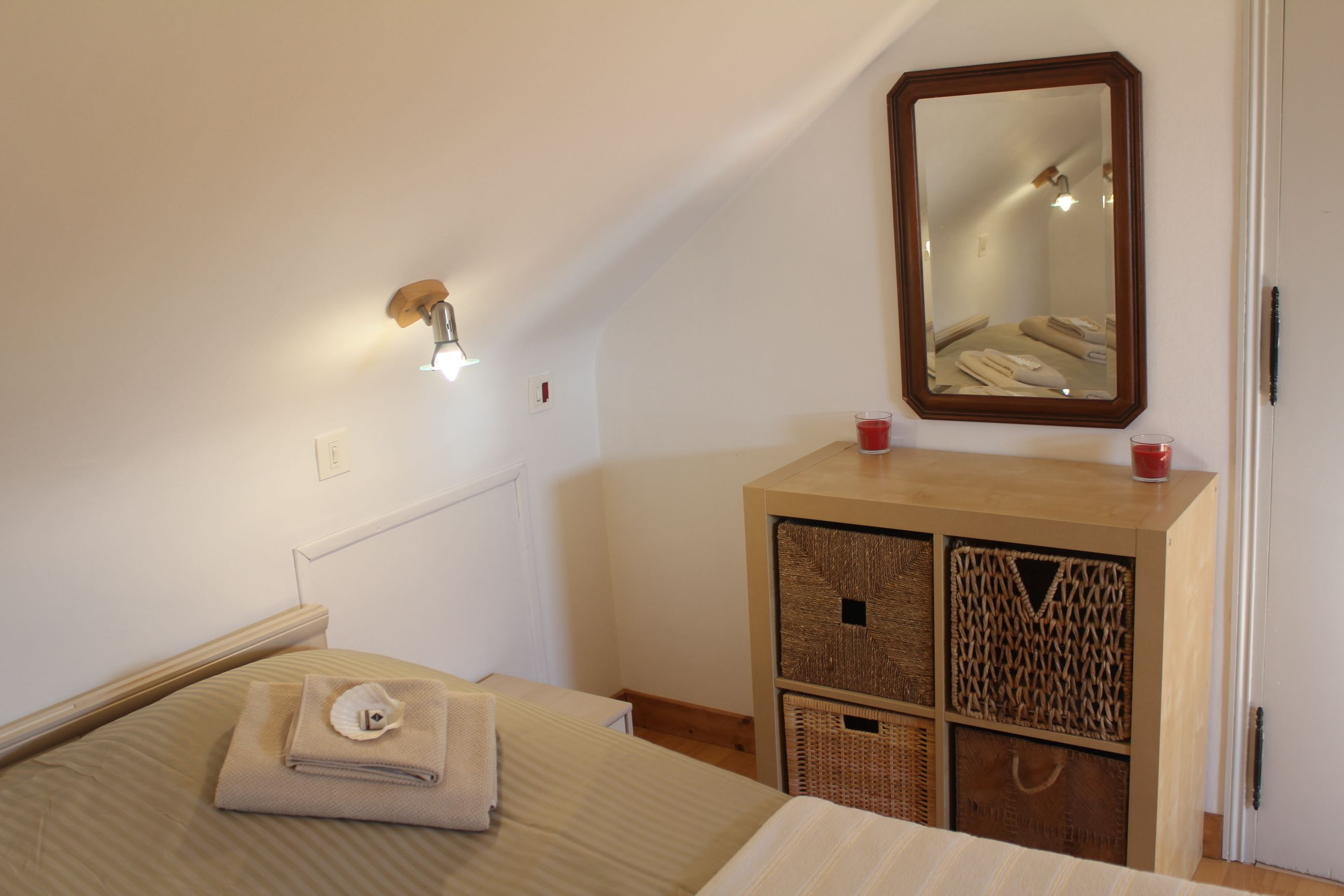 A bedroom with a bed, a bedside table with a lamp, a mirror, and a wooden cabinet with baskets.