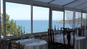 Een restaurant met gedekte tafels, grote ramen met uitzicht op zee en boten, en een kustlandschap.