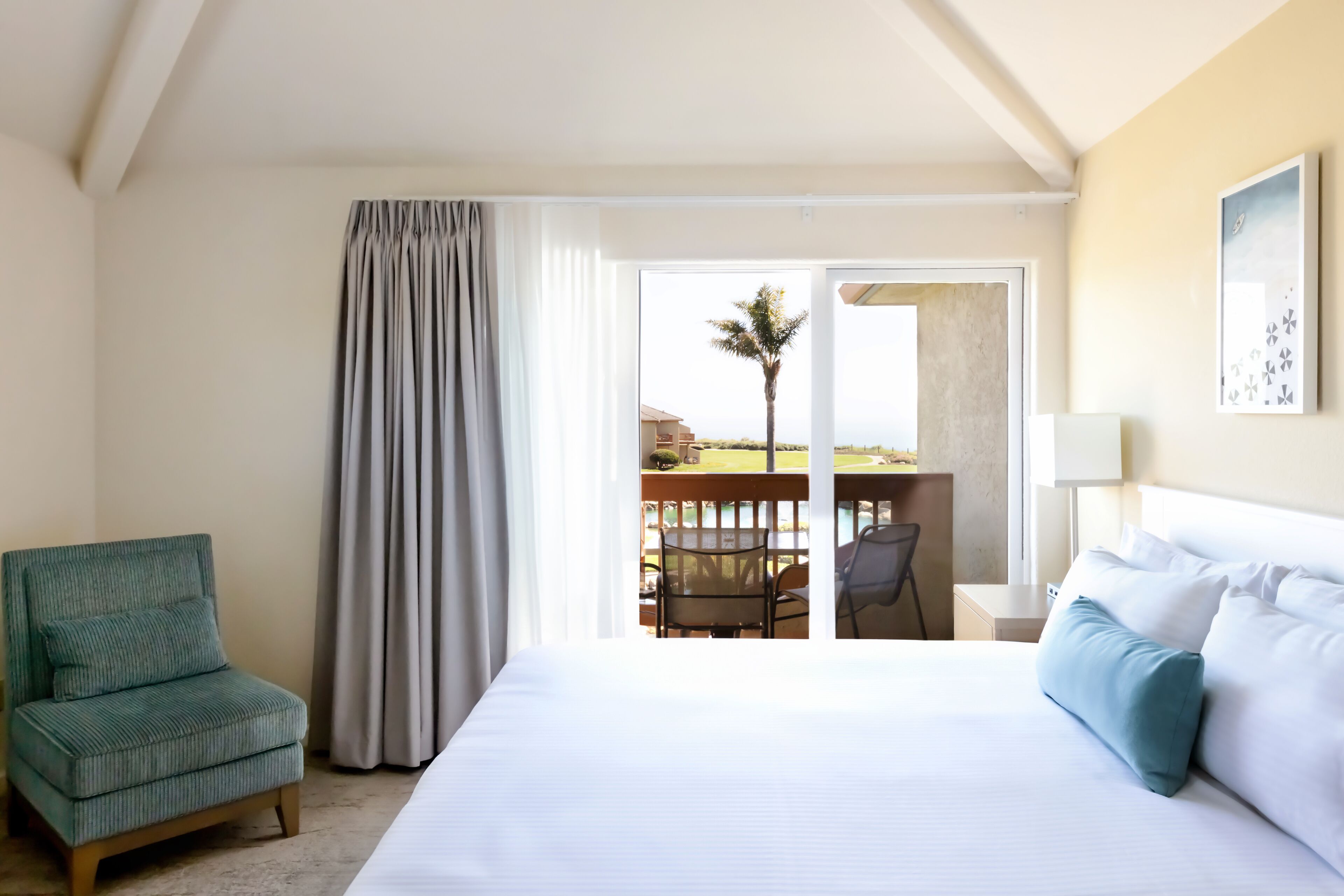 A hotel room with a bed, a green armchair, a balcony with a table and chairs, and a view of the outdoors.