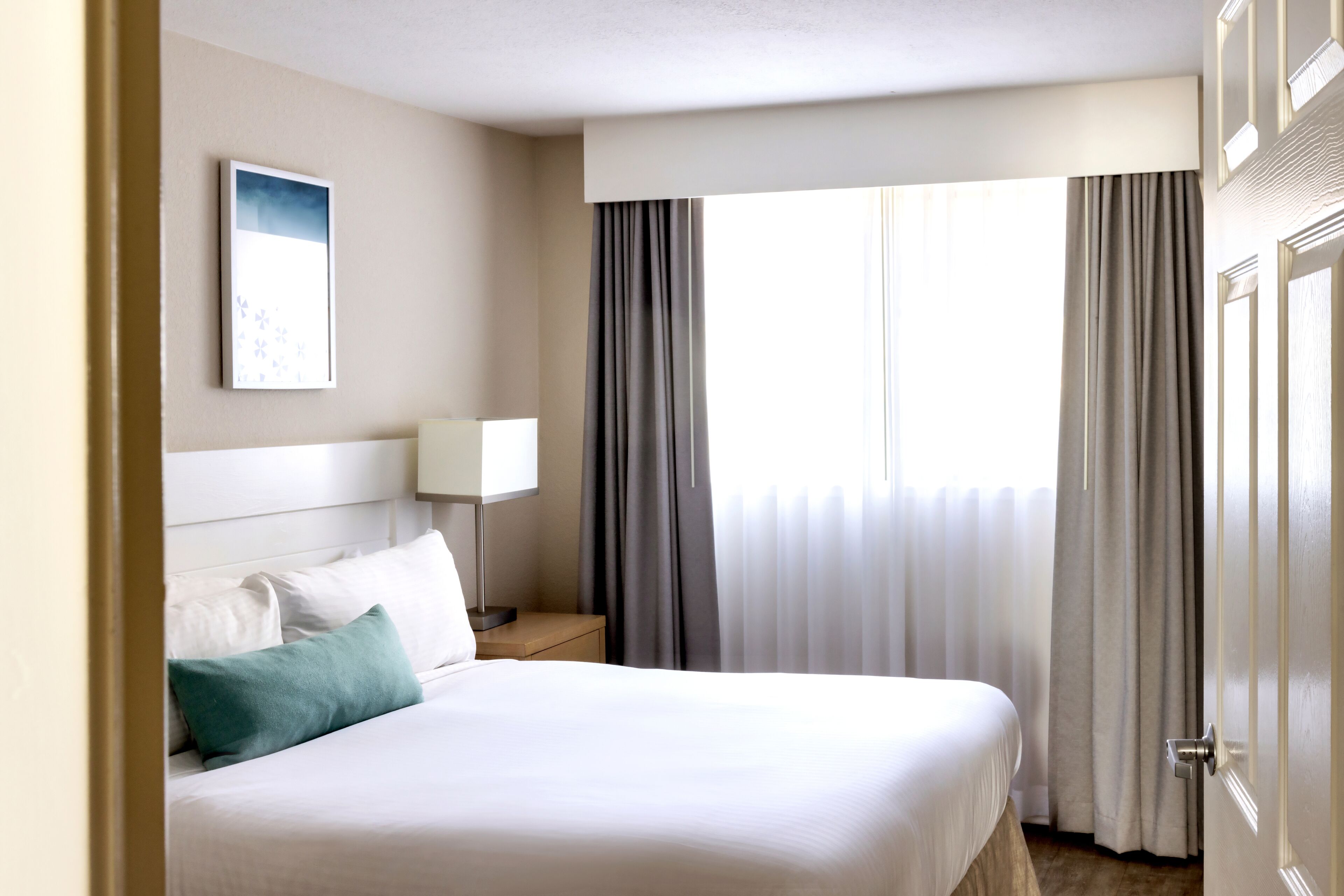 A hotel room with a large bed, a bedside table with a lamp, a window with curtains, and a framed picture on the wall.