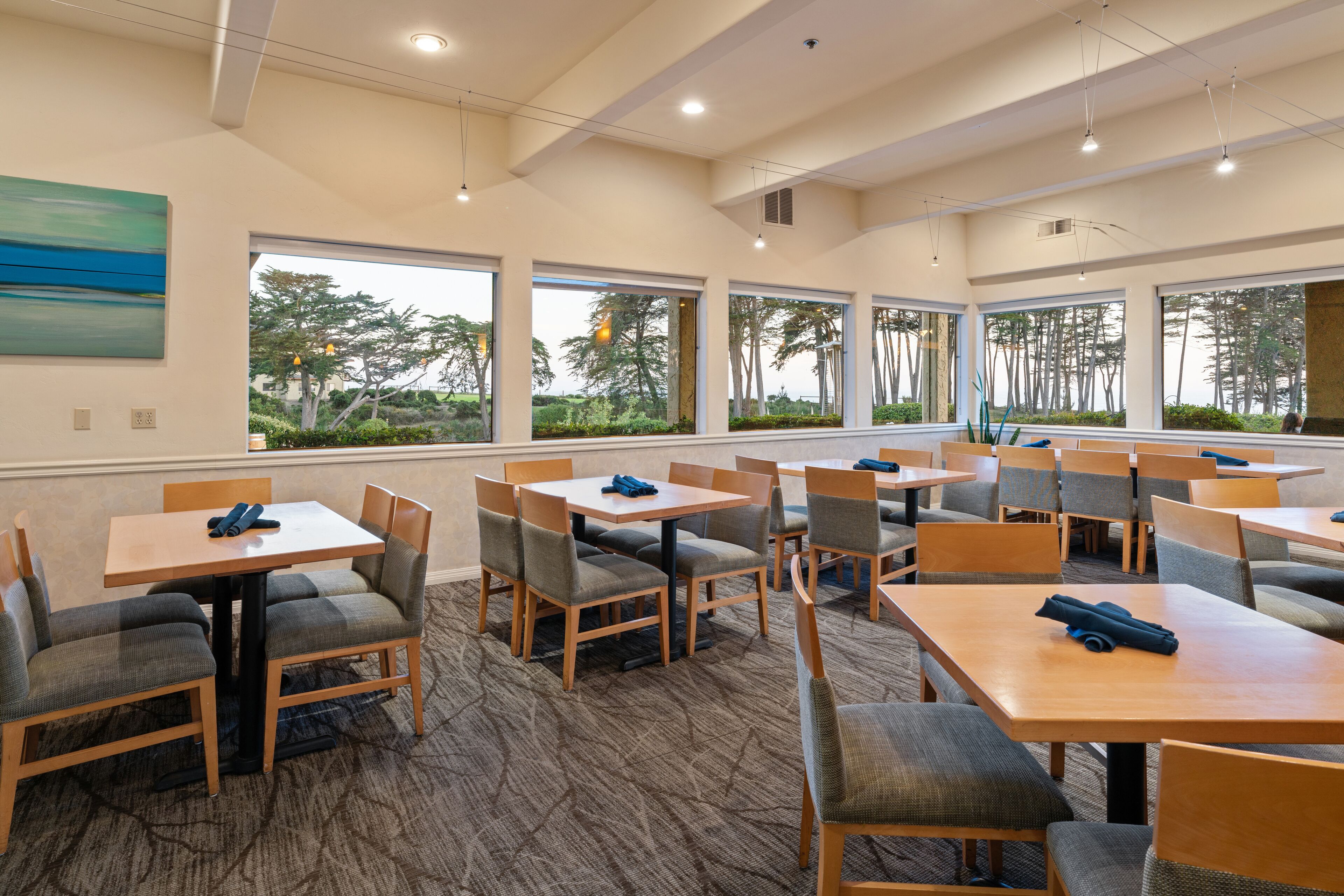 Une salle à manger d’hôtel avec de nombreuses tables et chaises, de grandes fenêtres donnant sur la verdure et un sol recouvert de moquette.