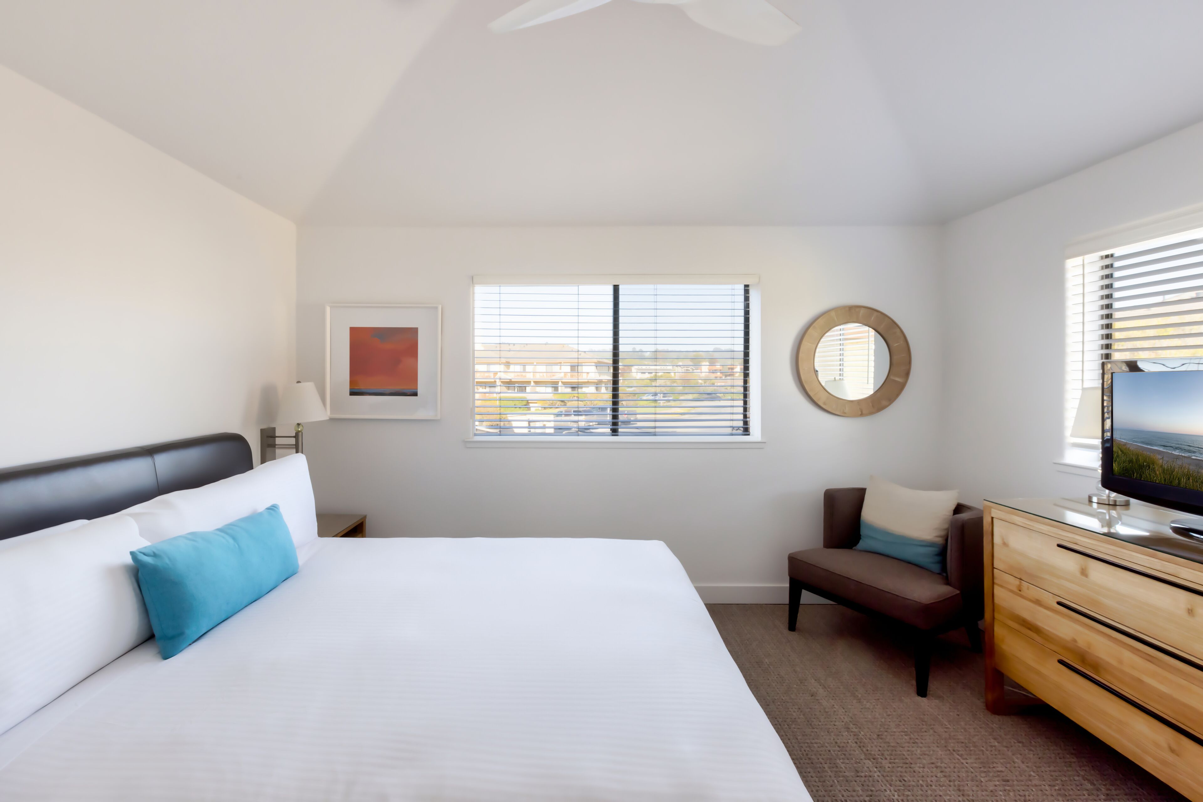 Quarto de hotel com cama, escrivaninha com televisão, cadeira, janela com persianas e ventoinha no teto.