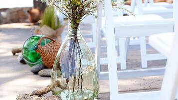 Un florero de cristal transparente con flores sobre una superficie de hormigón, rodeado de sillas blancas y un adorno verde.