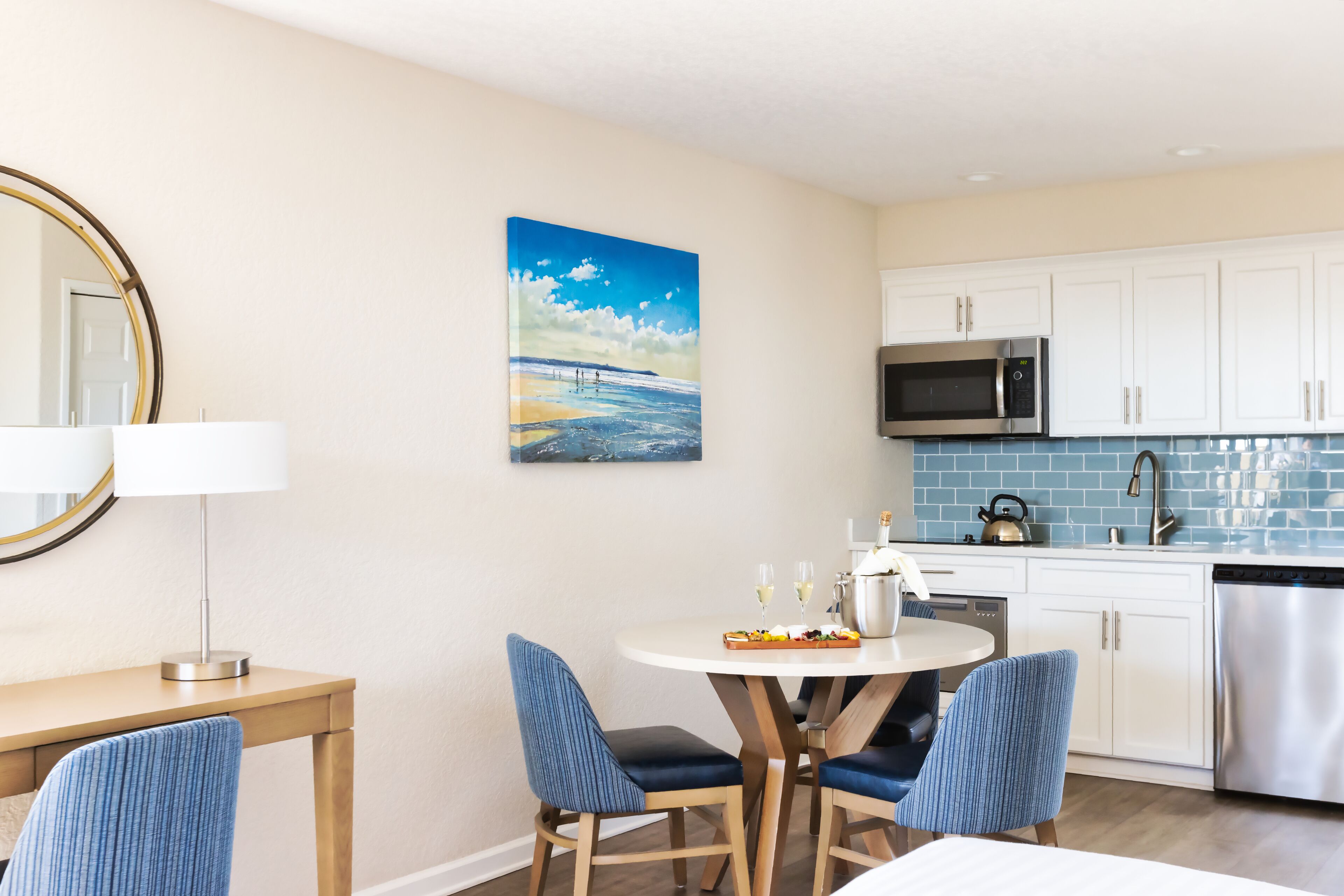 Una cocina moderna con armarios blancos, un microondas y un lavavajillas. Hay un comedor con una mesa y sillas, y un cuadro con una escena de playa en la pared.