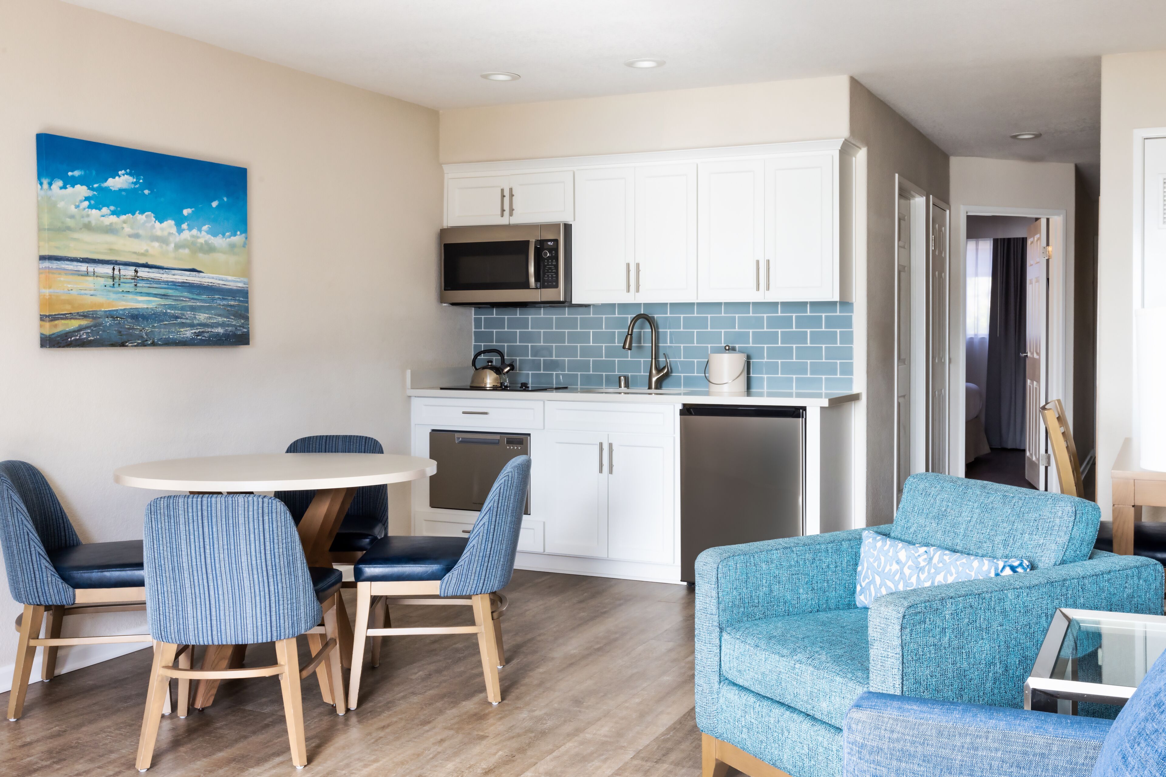 Een moderne keuken met witte kasten, een magnetron en een vaatwasser. Er is een eethoek met een ronde tafel en blauwe stoelen.