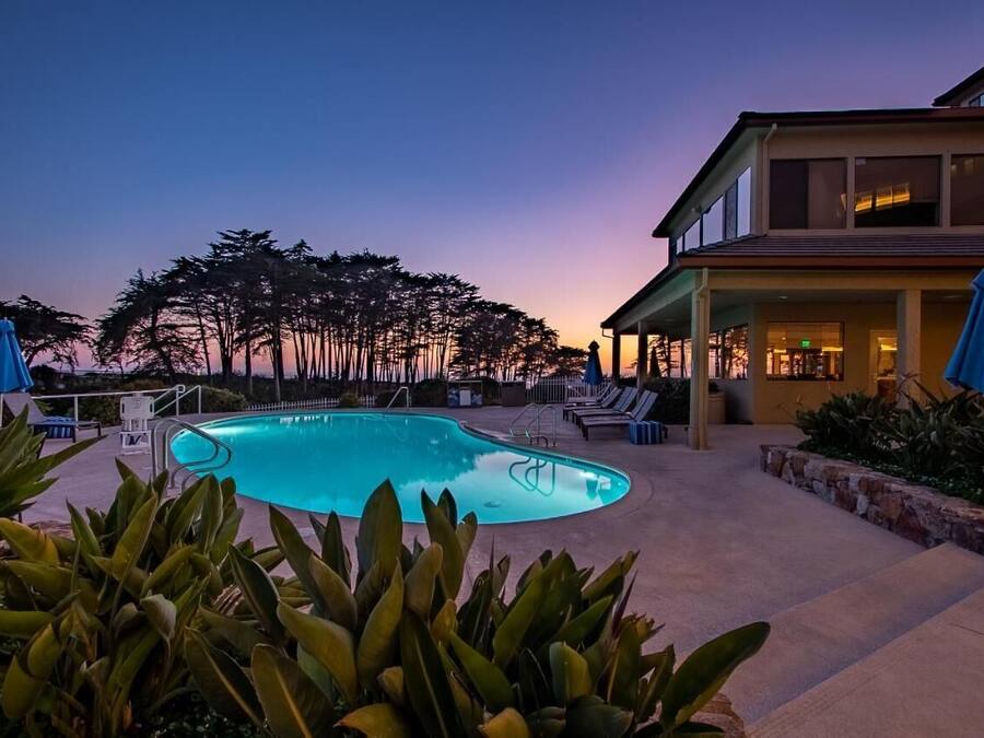 A swimming pool with lounge chairs and umbrellas, surrounded by a building with a covered patio area.