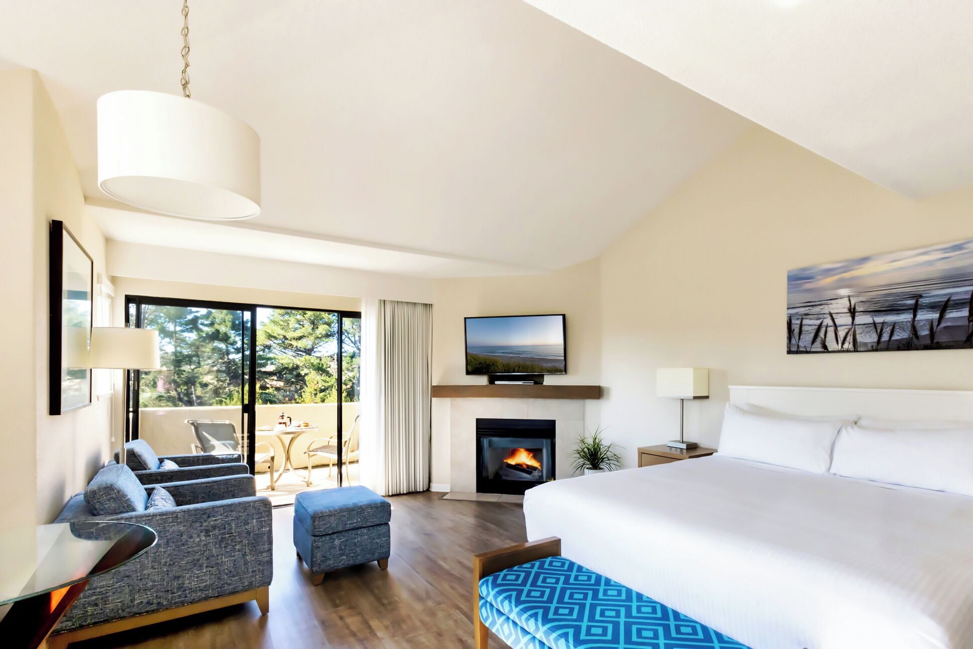 Una habitación de hotel moderna con una cama grande, un televisor de pantalla plana, una chimenea, un balcón con vista a árboles y un área de comedor con una mesa y sillas.