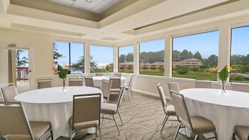 Un salón de conferencias con mesas y sillas redondas, ventanas amplias con vistas a edificios y zonas verdes.
