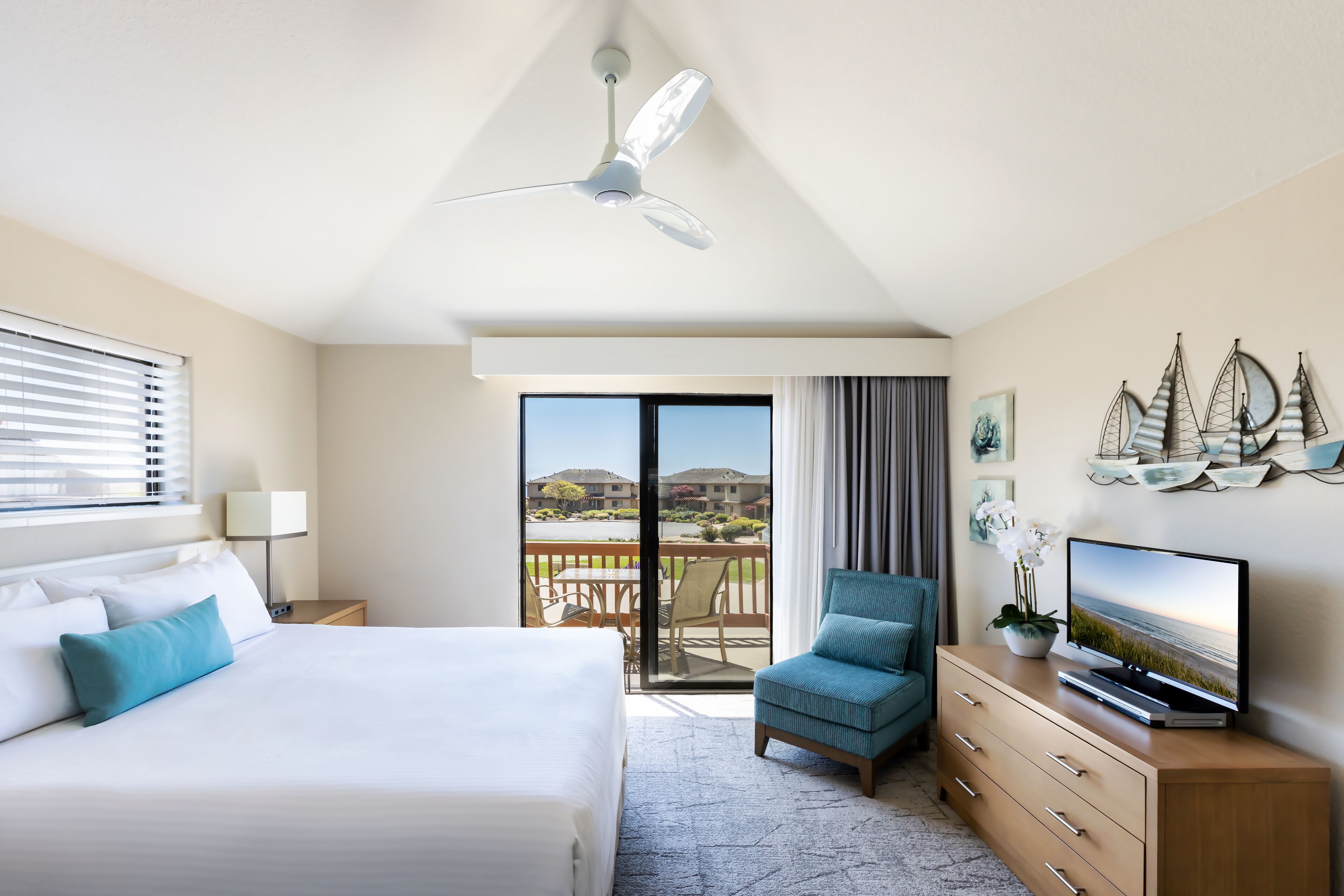 A hotel room with a bed, a chair, a TV, a ceiling fan, and a balcony with a view.