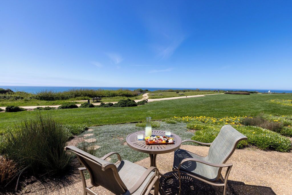 A patio with two chairs and a table, overlooking a grassy area with a path and distant view of the ocean.
