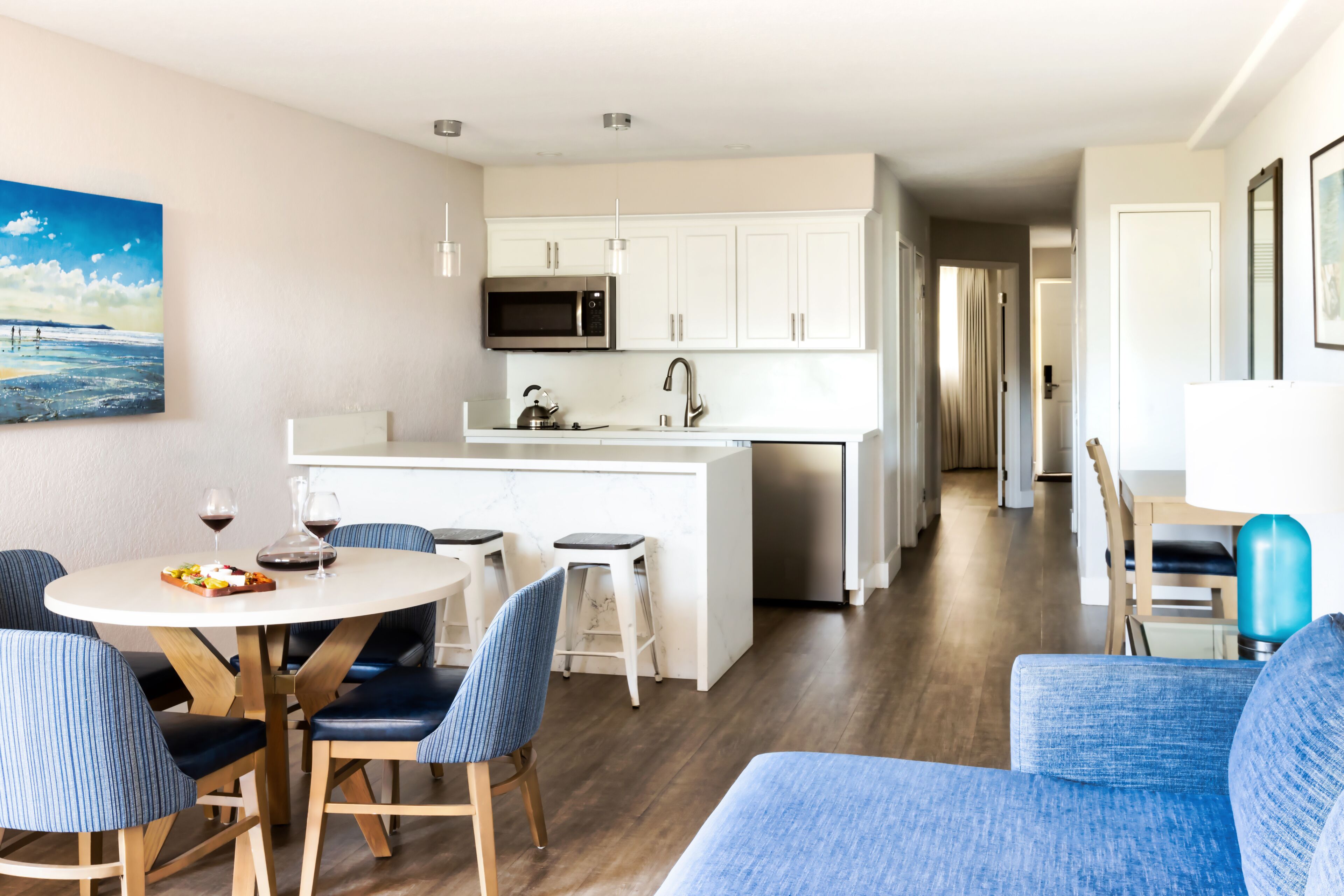 Una cocina moderna con armarios blancos, microondas y pileta de acero inoxidable. Hay un comedor con una mesa redonda y sillas azules.