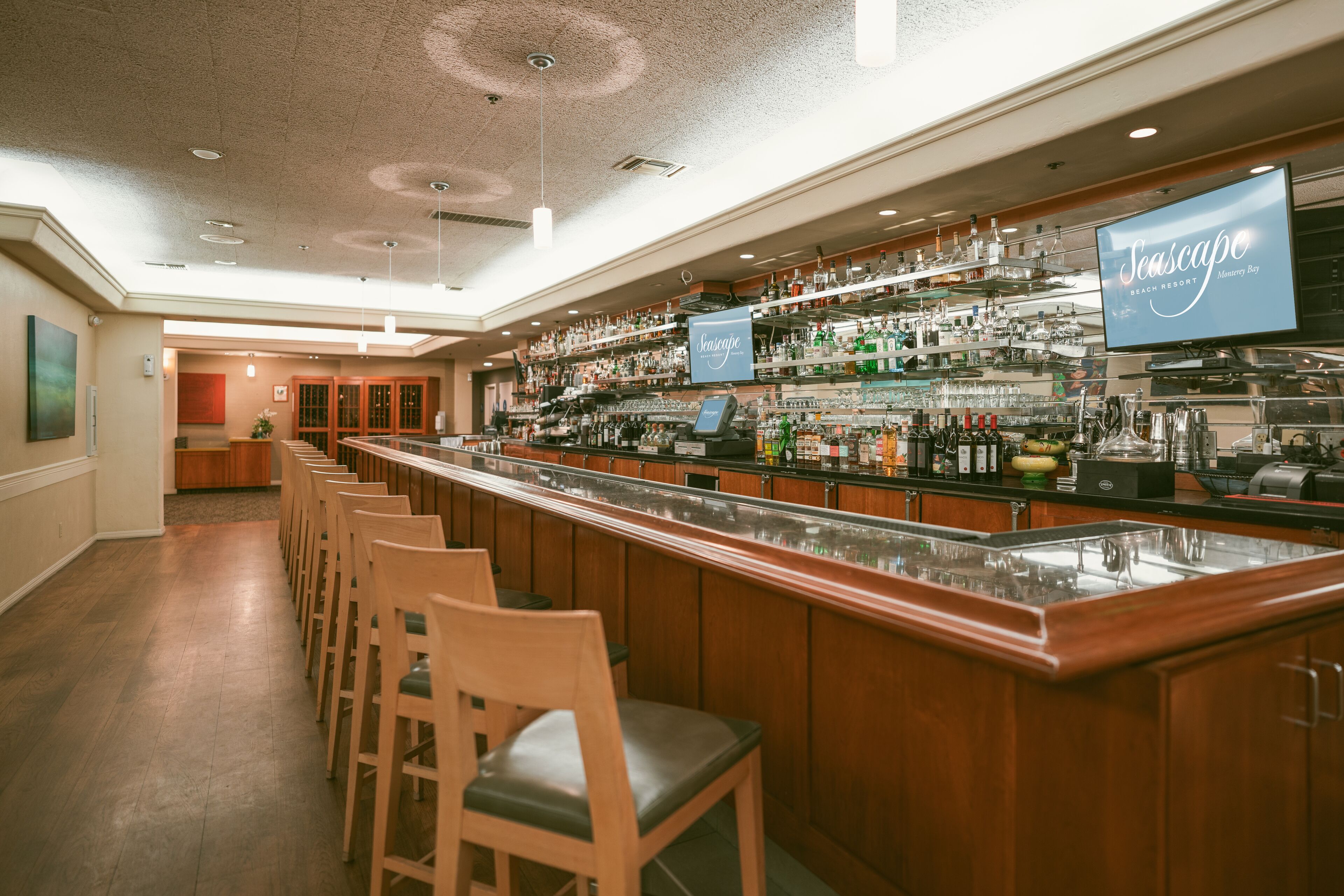 Una zona de bar con sillas de madera, una barra larga y estanterías llenas de botellas y vasos.