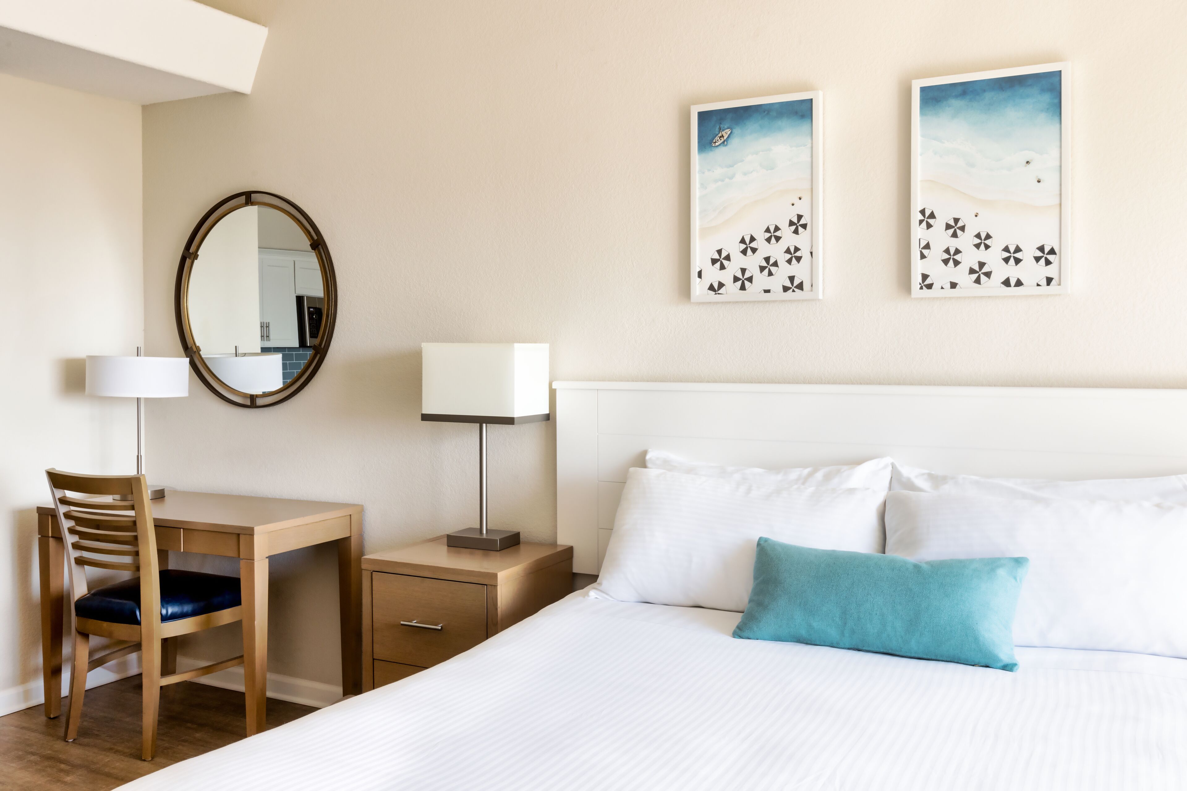 A hotel room with a bed, a desk with a chair, a nightstand with a lamp, a mirror, and two wall art pieces.