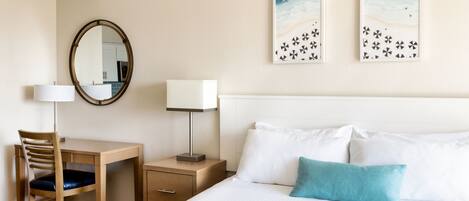 A hotel room with a bed, a desk with a chair, a nightstand with a lamp, a mirror, and two wall art pieces.