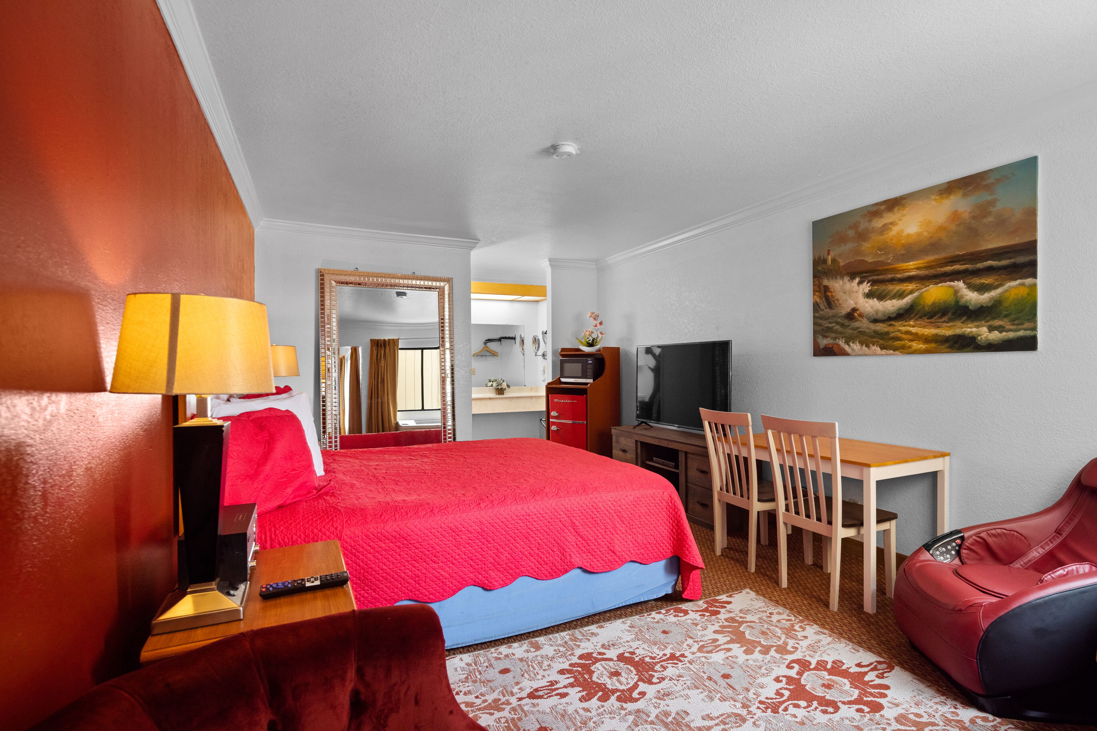 Une chambre d’hôtel avec un lit rouge, un bureau, une chaise, un fauteuil de massage, une télévision et un tableau représentant un paysage marin.