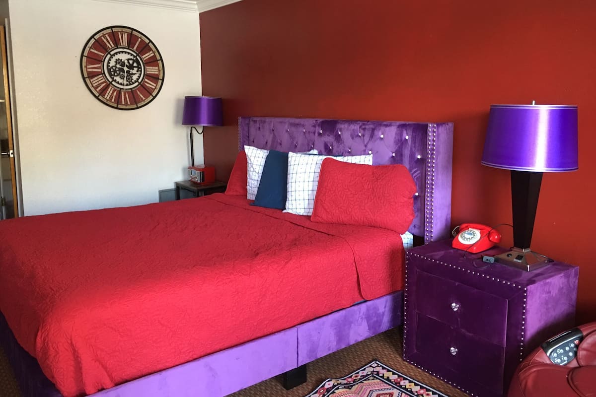 A bedroom with a red bedspread, a purple headboard, and a clock on the wall.
