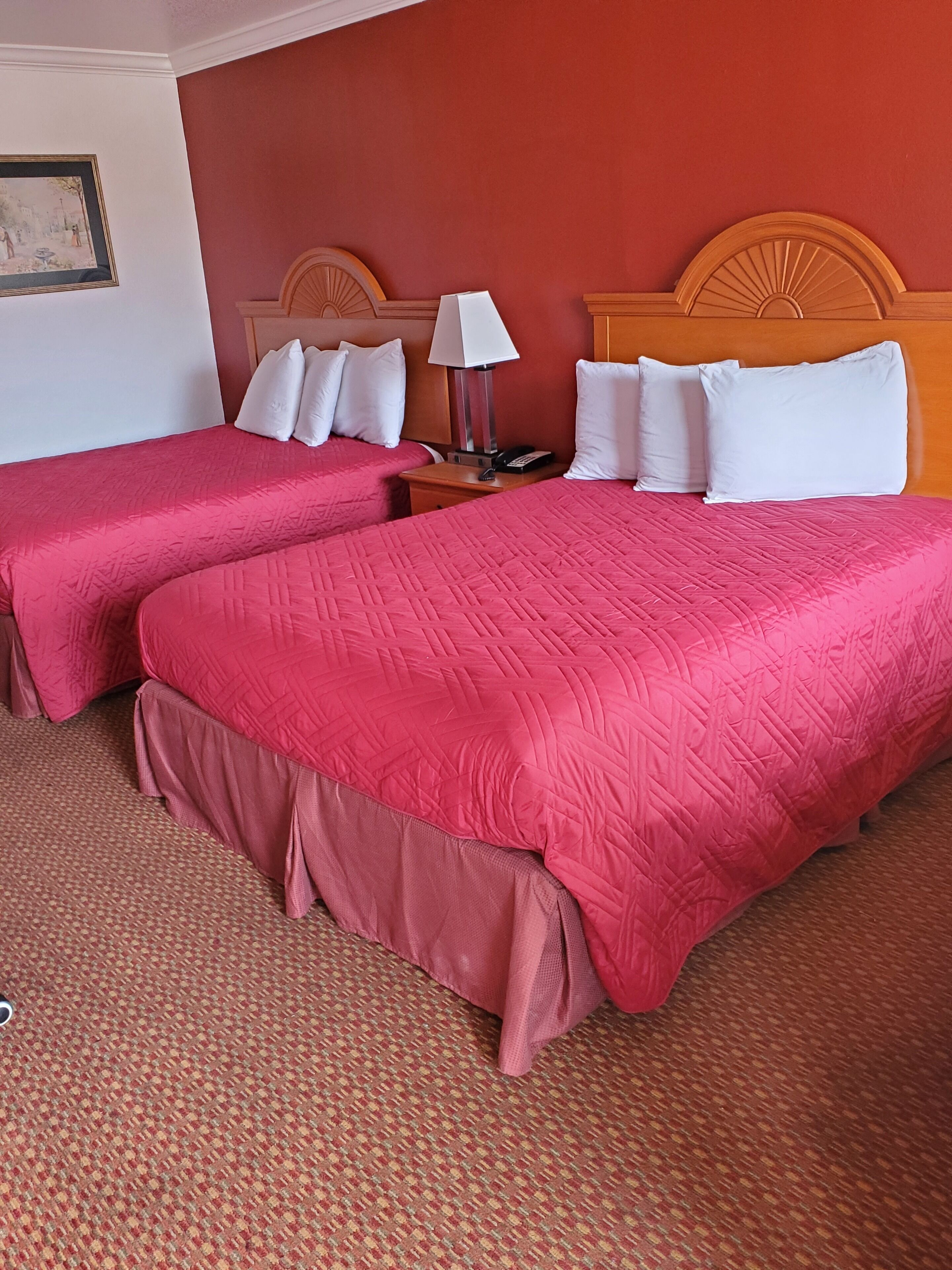 Een hotelkamer met twee bedden, elk voorzien van een roze sprei en witte kussens. Er is een houten hoofdbord, een nachtkastje met een lamp en een lijst met een afbeelding aan de muur.