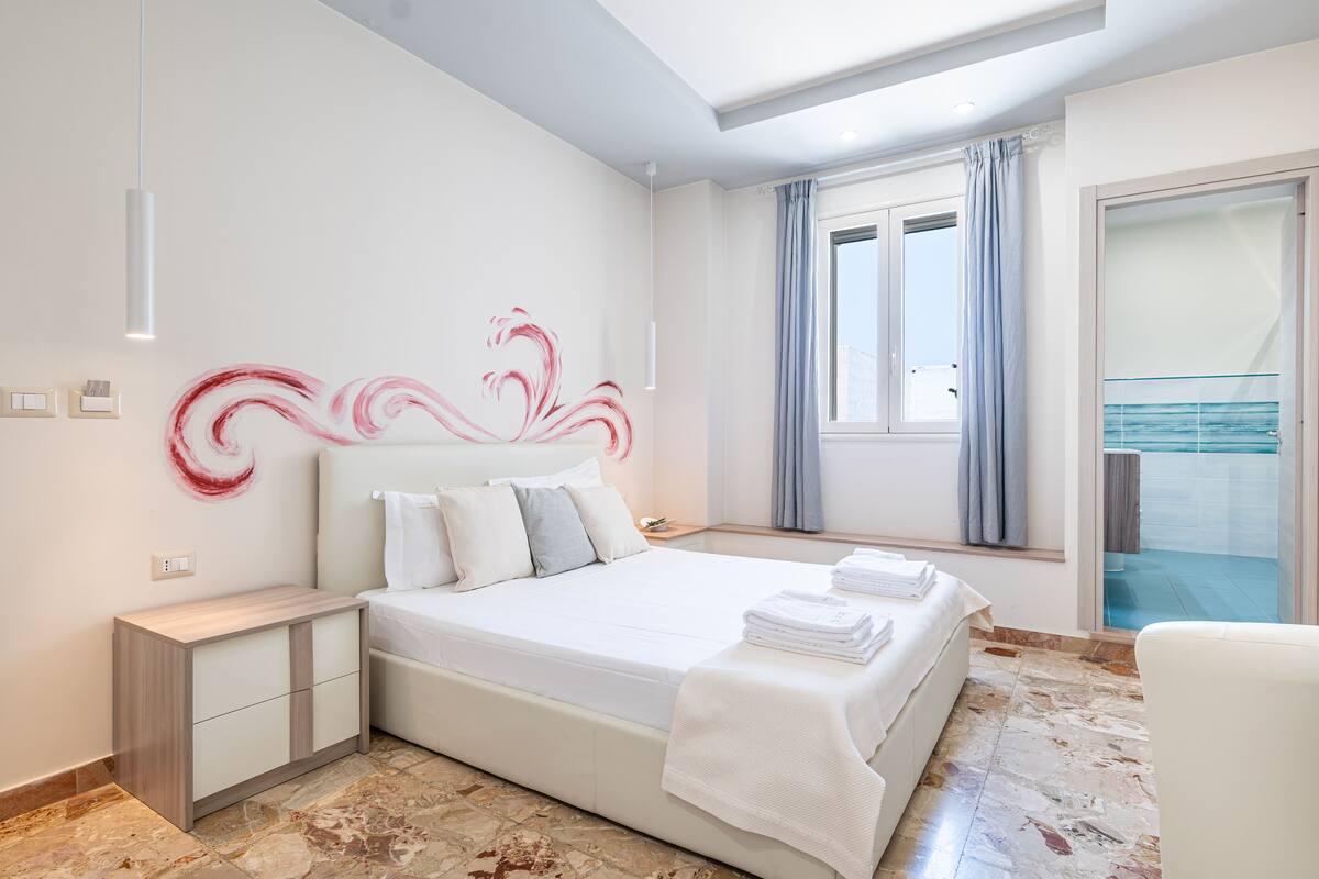 Un dormitorio moderno con una cama grande, una mesita de noche, una ventana con cortinas y vista al mar.