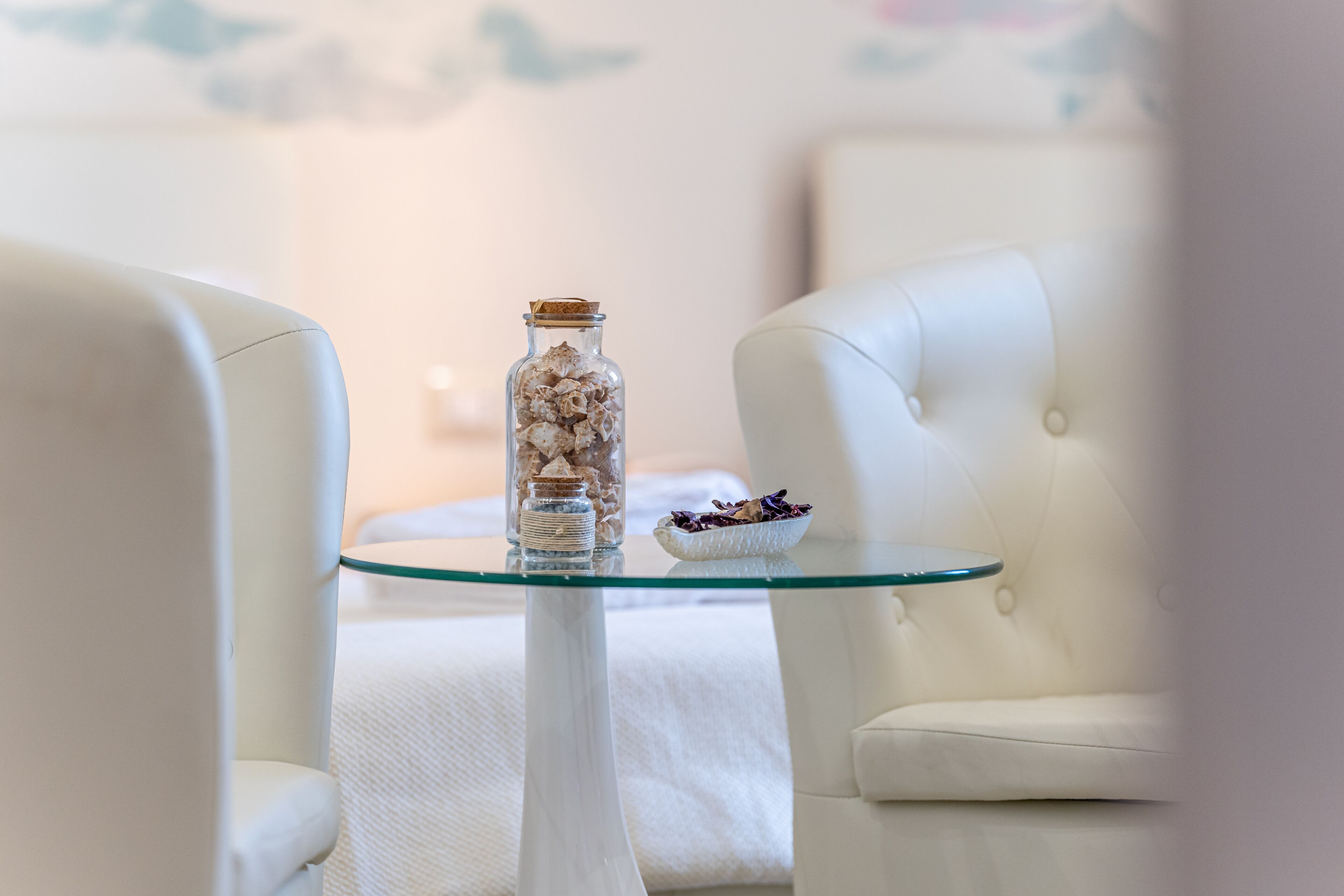 A room with a glass-top table, a white vase with dried flowers, and a bowl of purple flowers.