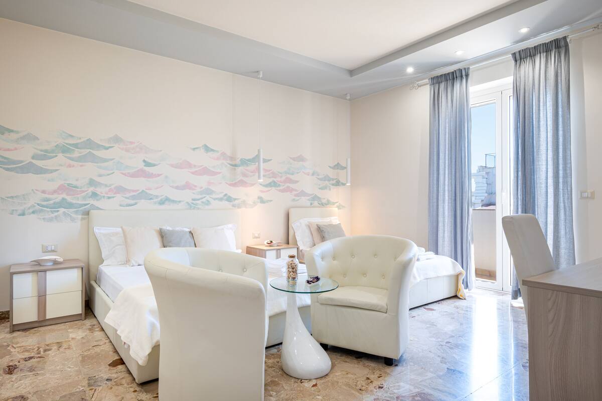 Una habitación de hotel moderna con una cama, dos sillones, una mesa de centro de cristal y vistas a la ciudad a través de grandes ventanales.