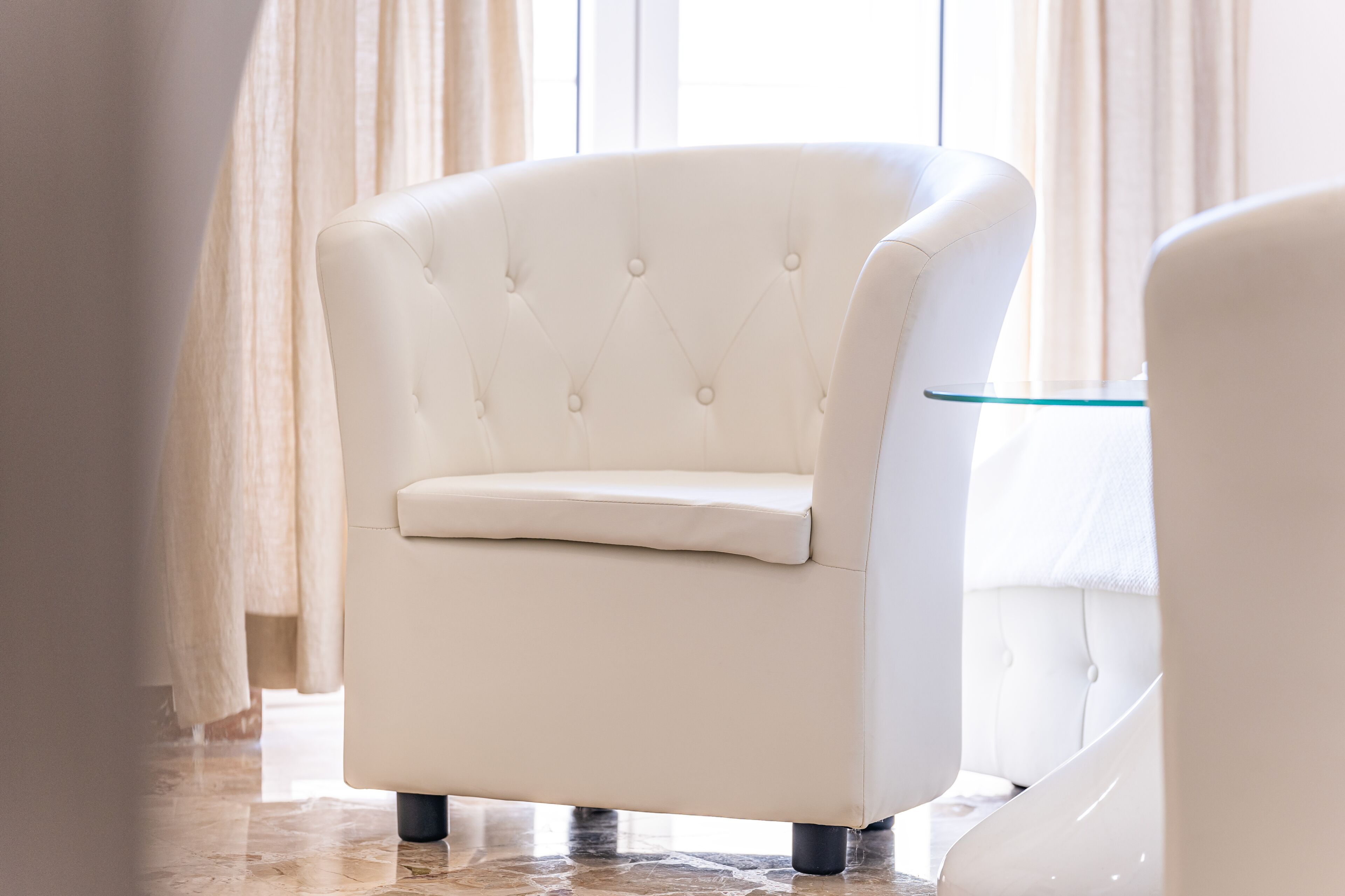 A white armchair with a cushioned seat and backrest, positioned in a room with a glass table and curtains.