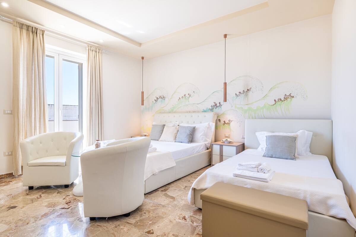 Habitación de hotel con dos camas, un área de descanso con una silla, una mesita y un gran ventanal con cortinas.