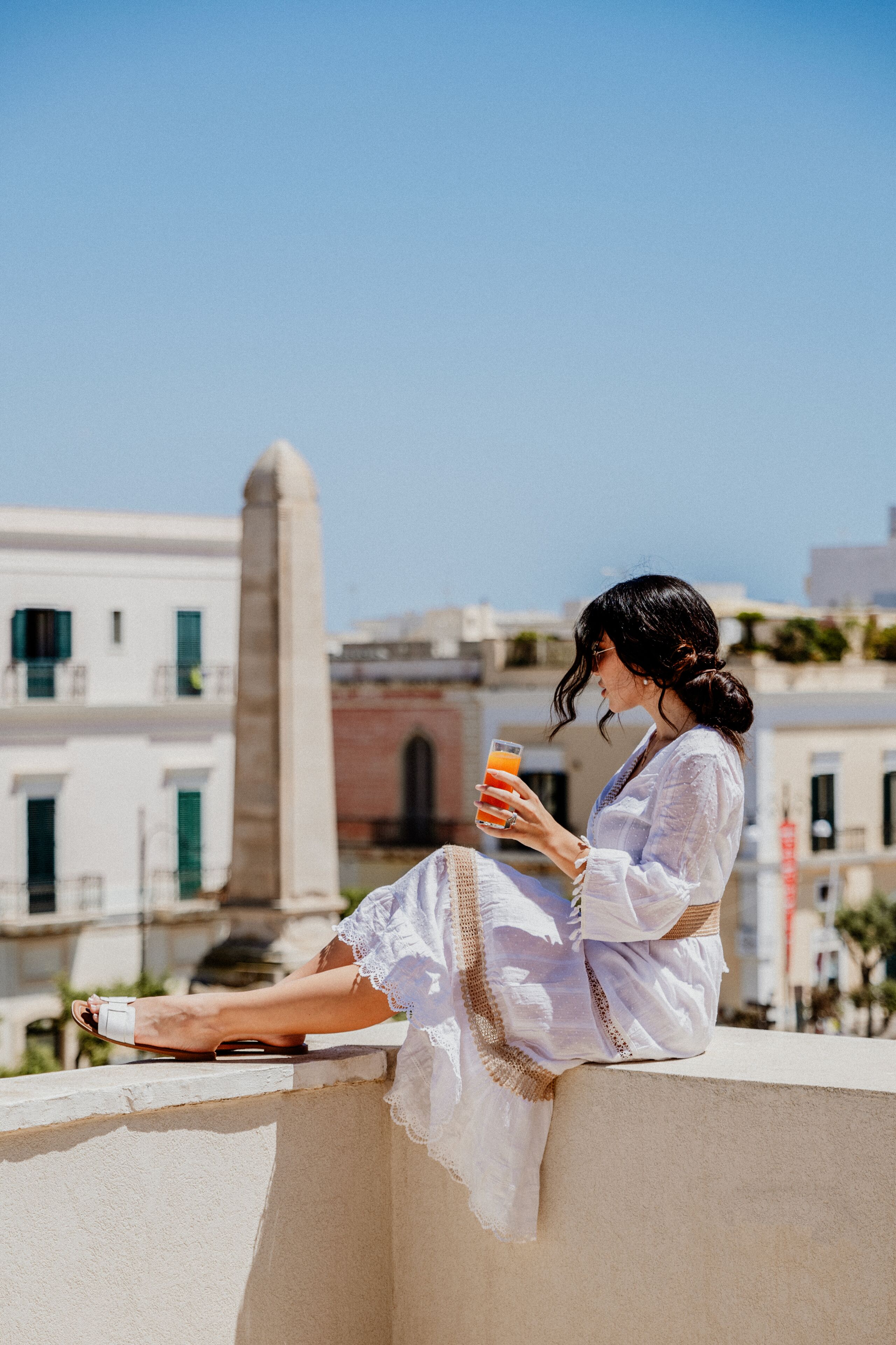 A person sitting on a balcony with a drink, overlooking a cityscape with buildings and a tall monument.