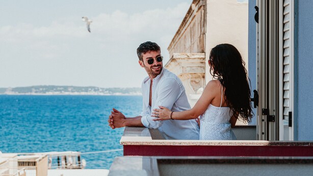 Una pareja en un balcón con vista al mar, con un edificio y un ave al fondo.