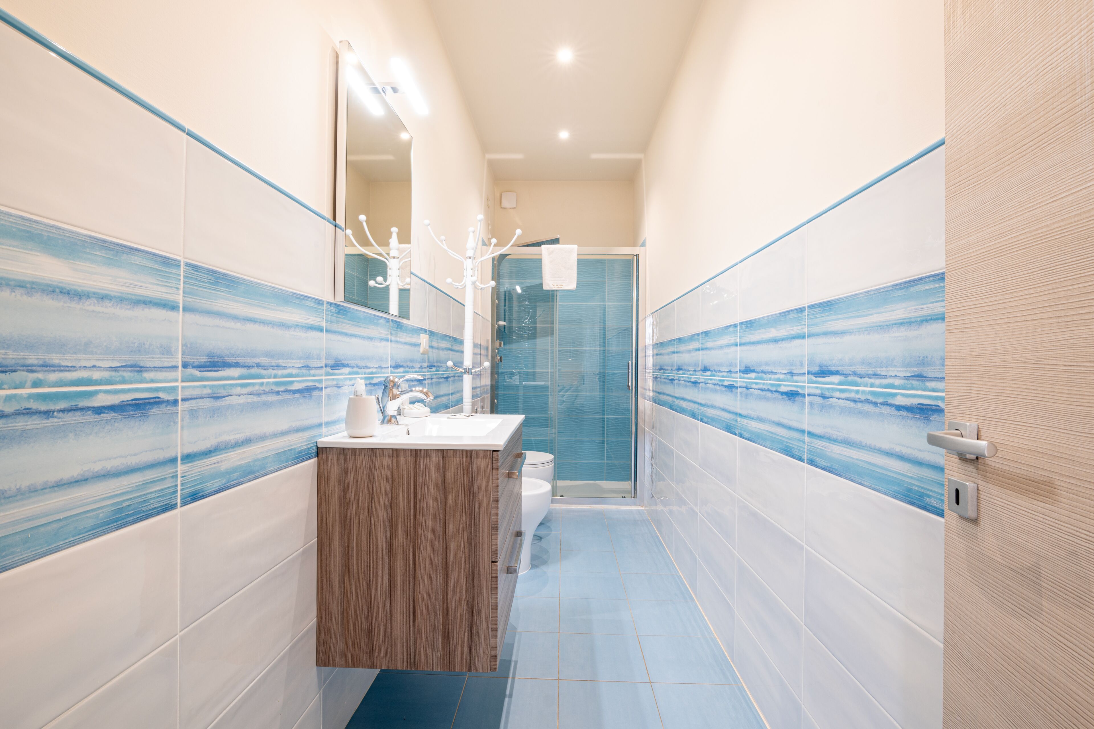 A narrow bathroom with a wooden vanity, a glass shower enclosure, and a tiled wall with a blue wave pattern.