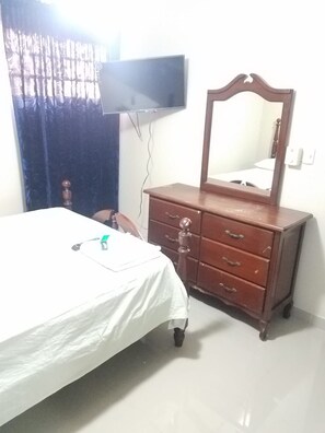 Ein Schlafzimmer mit einem Bett, einem Schminktisch mit Spiegel, einem an der Wand befestigten Fernseher und einem Vorhang.