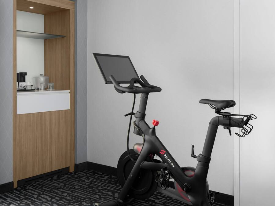 A modern hotel room with a stationary bike, a flat-screen TV, and a small kitchenette area.