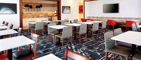 A hotel dining area with multiple tables and chairs, a kitchen in the background, and a flat-screen TV mounted on the wall.