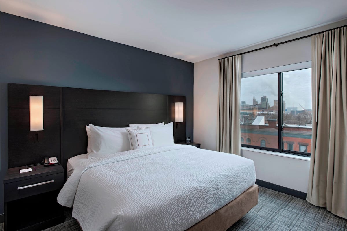 Une chambre d’hôtel avec un grand lit, une tête de lit sombre, des lampes de chevet, une table de chevet et une fenêtre avec des rideaux.