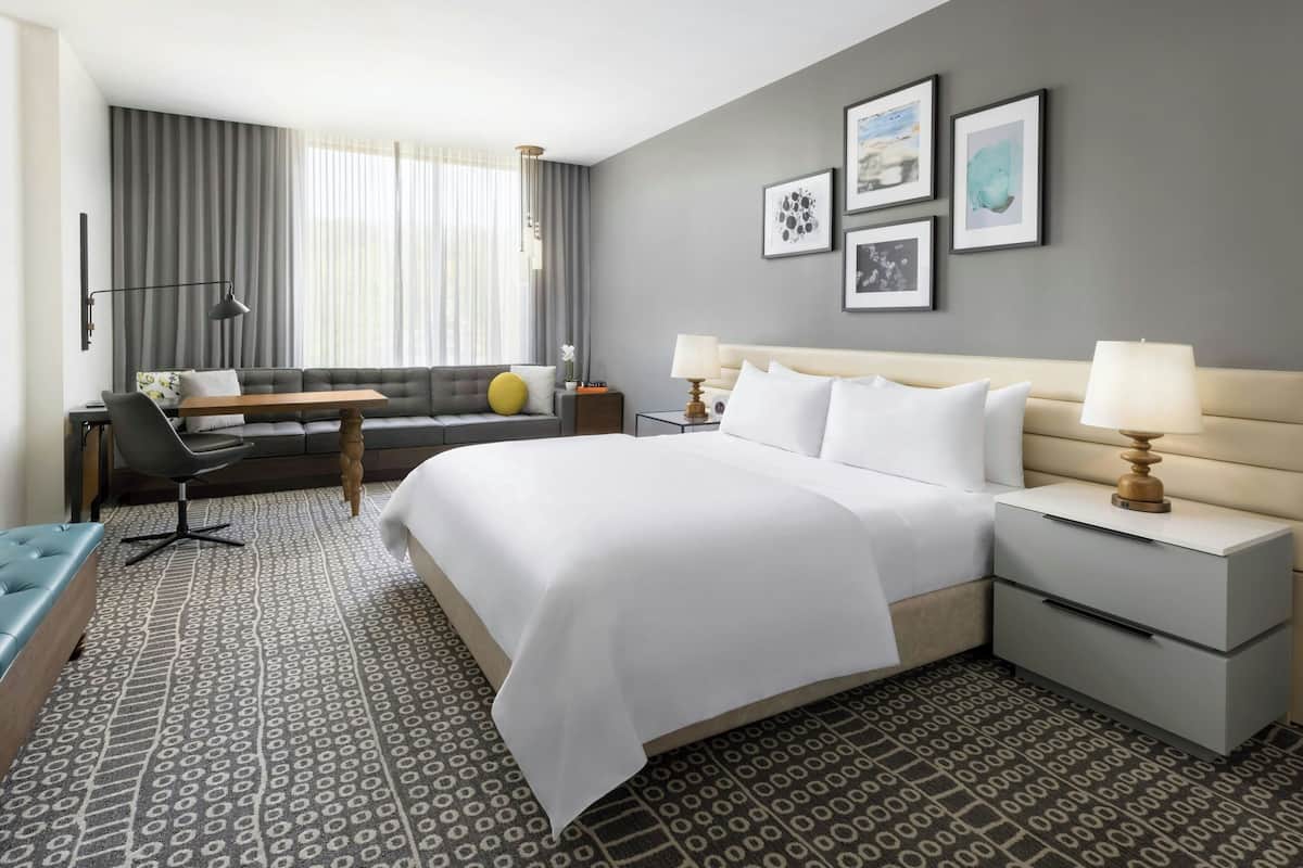Habitación de hotel con una cama grande, un sofá, un escritorio y una silla. Hay cuadros en la pared y una ventana con cortinas.