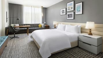 Hotelkamer met een groot bed, een bank, een bureau en een stoel. Er hangen foto’s in lijsten aan de muur en een raam met gordijnen.