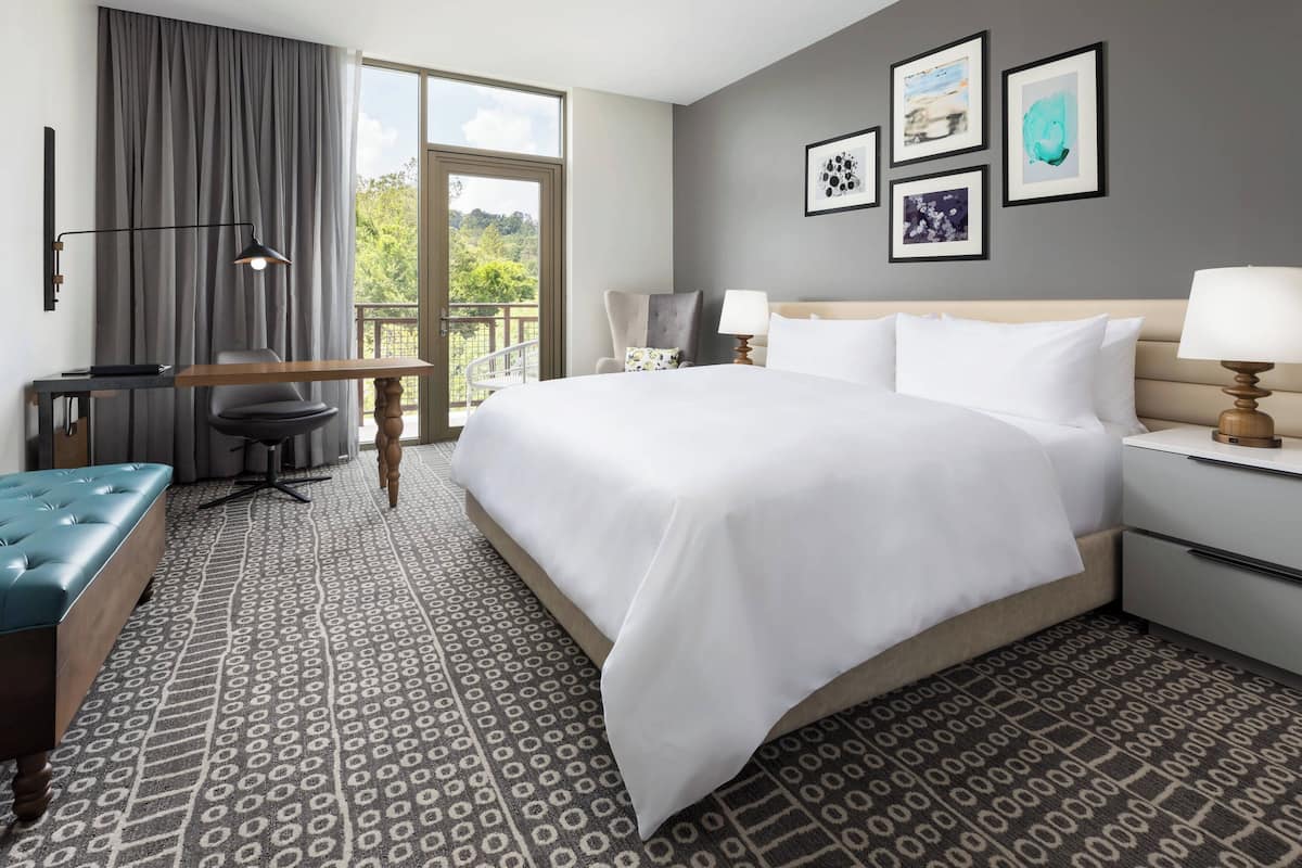 Habitación de hotel con una cama grande, un escritorio, una silla, una mesita y un balcón con vistas a zonas verdes.