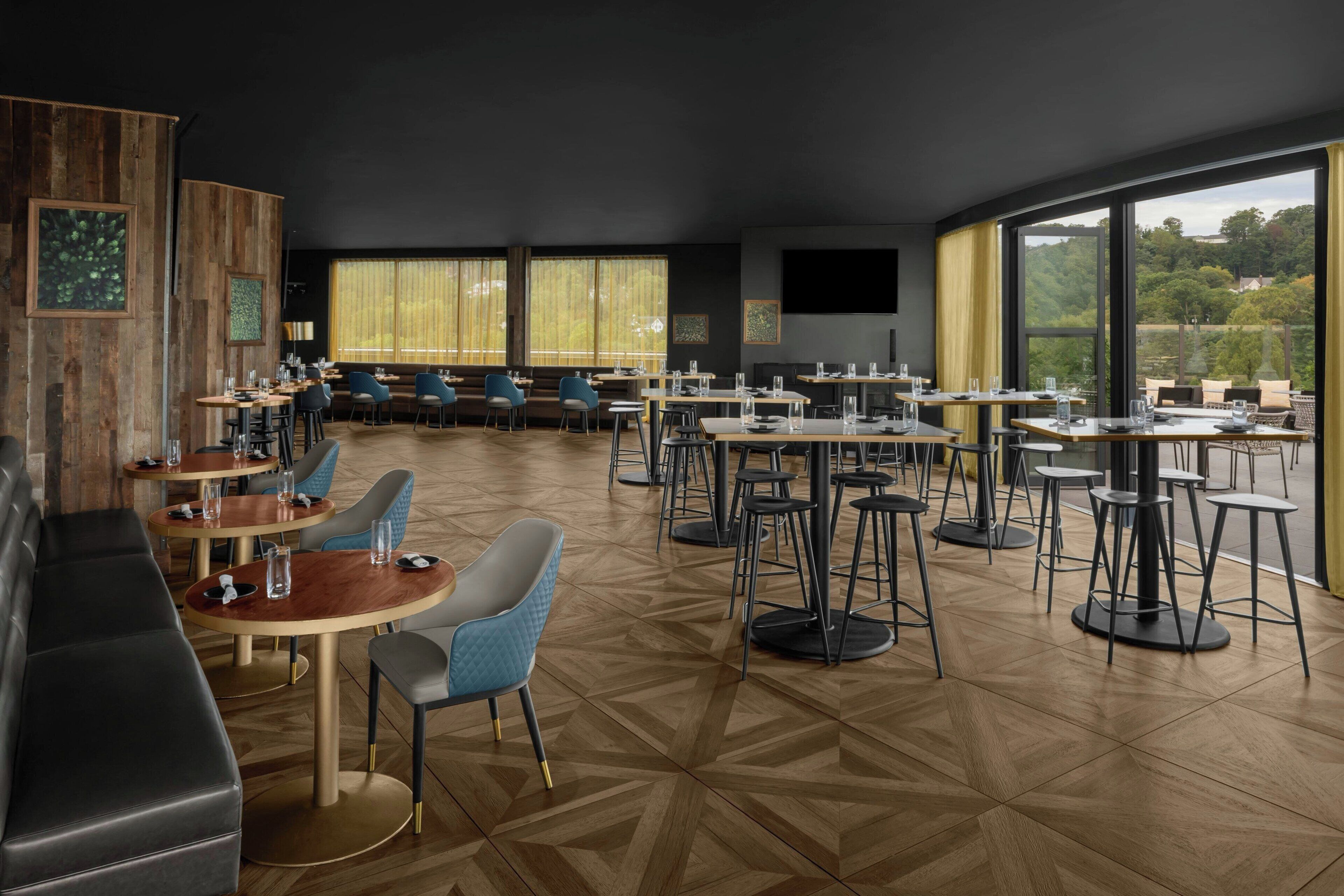 A modern restaurant with a mix of booths and tables, large windows offering a view of the outdoors, and a wooden floor with a geometric pattern.