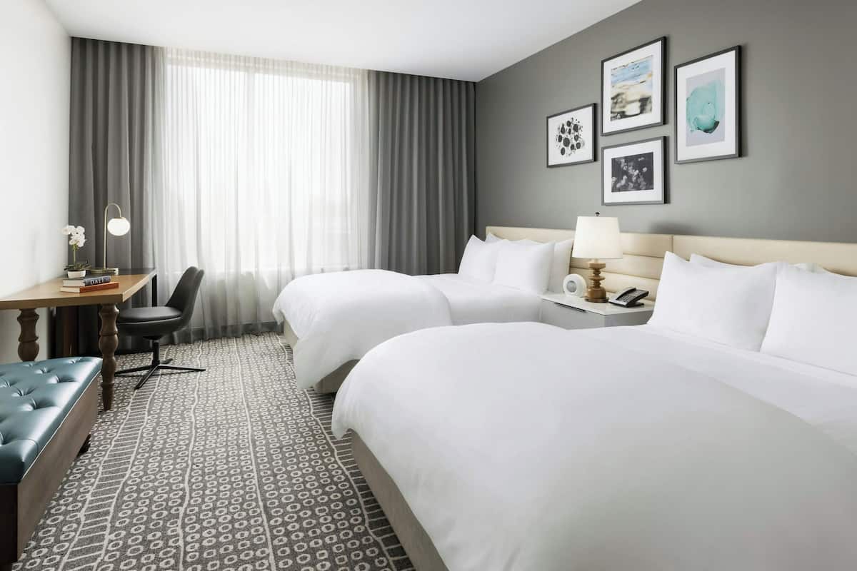 Habitación de hotel con dos camas, un escritorio con silla, una lámpara y cuadros en la pared.