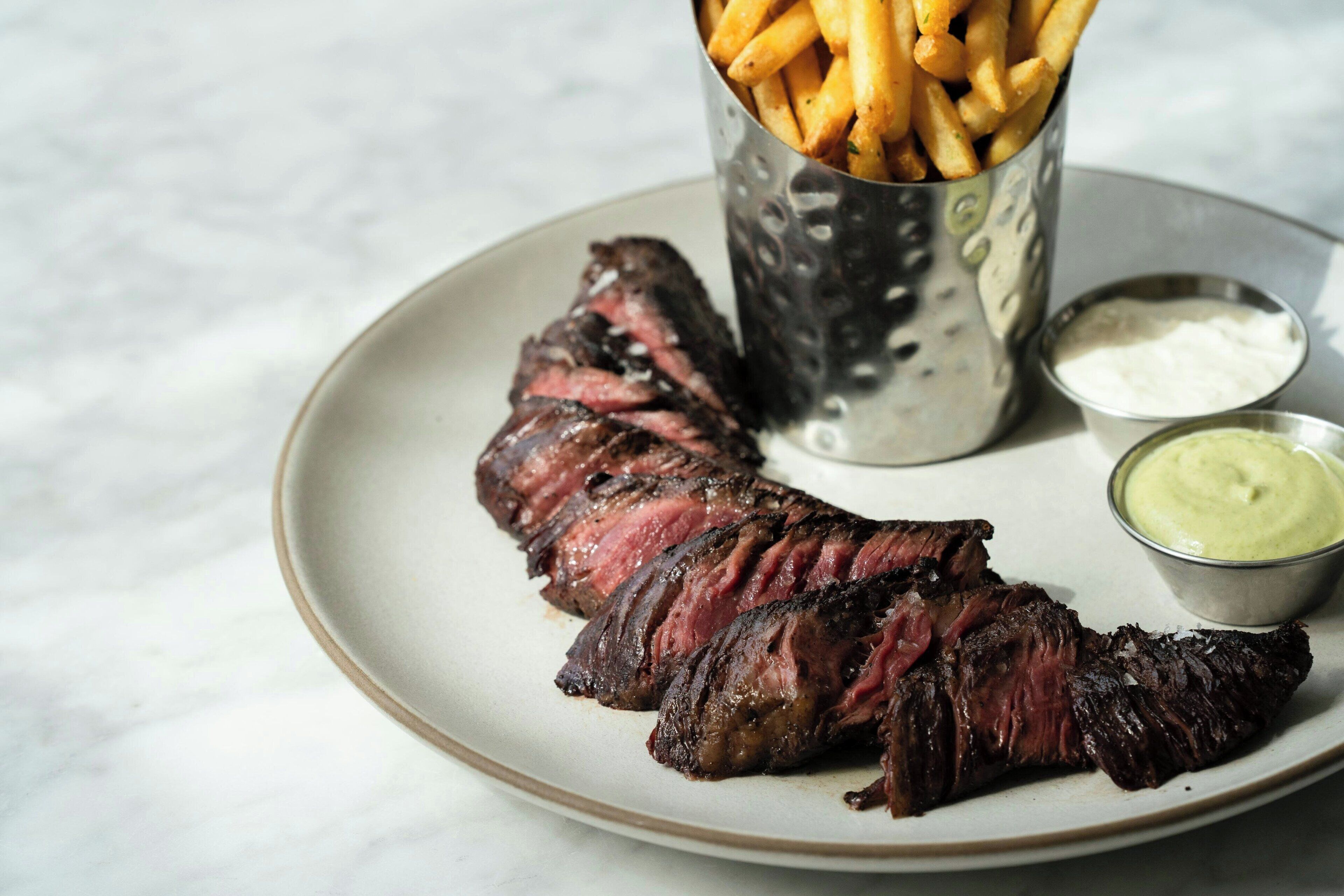 Een bord met plakjes steak, friet en twee dipsausen.