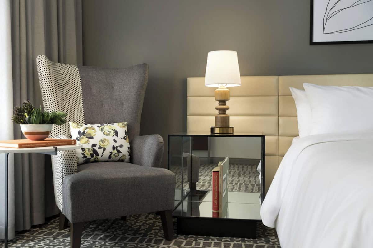Habitación de hotel con un sillón gris, una mesita de noche con lámpara, cama con ropa blanca y una obra de arte enmarcada en la pared.