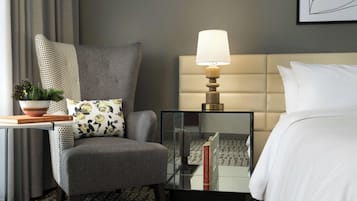 Habitación de hotel con un sillón gris, una mesita de noche con lámpara, cama con ropa blanca y una obra de arte enmarcada en la pared.